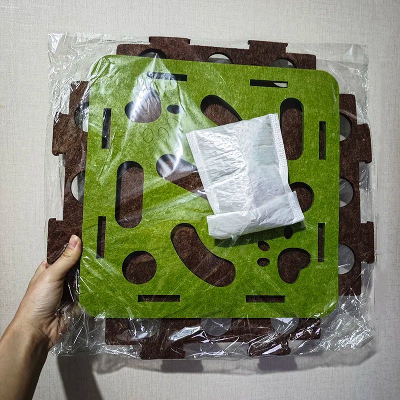 Green and brown puzzle mat with a white square object on top, held by a hand against a light gray background.