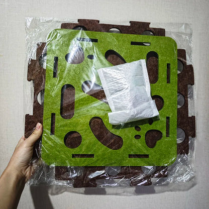 Green and brown puzzle mat with a white square object on top, held by a hand against a light gray background.